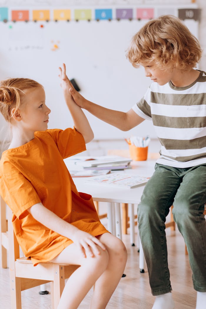 Two children high five while sitting in a bright classroom, promoting friendship and teamwork.
