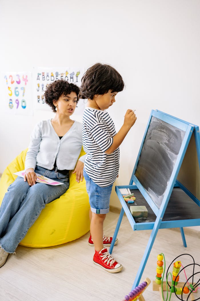 A young boy writes on a blackboard with a teacher observing, creating an educational moment.
